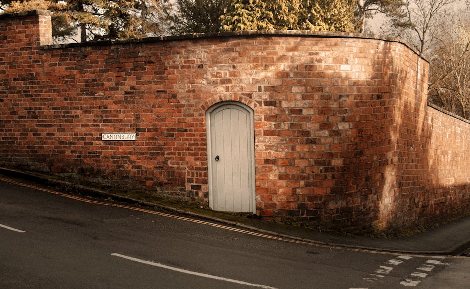 A curved brick building with reddish-brown bricks forming a boundary wall along a quiet street. The wall features a small, arched white door situated near the centre and a black door handle. To the left of the door, there is a white rectangular street sign reading 'CANONBURY' attached to the brickwork. The building is set slightly above street level, with a narrow pavement and a gentle slope leading up to the door. The street surface is dark asphalt with white road markings, and there are areas of moss or dirt near the base of the wall. In the background, leafless trees and a clear sky are visible, suggesting a late autumn or winter setting. The scene is illuminated by natural light, with shadows cast on the brick wall. This setting could relate to house removals or furniture transport, highlighting the accessibility considerations for home relocation services provided by Removal Companies Watford, especially with narrow road access involved.