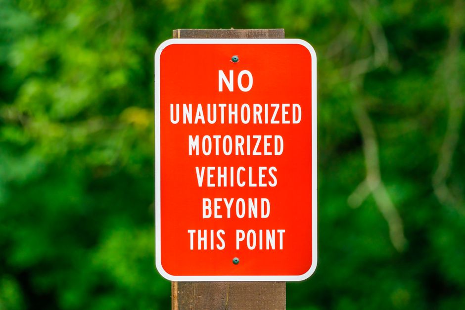 A rectangular red and white traffic sign mounted on a wooden post outdoors, indicating that no unauthorized motorized vehicles are allowed beyond that point. The sign features bold white text reading 'NO UNAUTHORIZED MOTORIZED VEHICLES BEYOND THIS POINT' on a red background, with a white border around the edges. The background of the image shows out-of-focus greenery, suggesting a natural setting such as a park or residential area with trees. This type of signage is common in environments where access is restricted for vehicles, and during home relocation or furniture transport, such signage can assist in guiding vehicle routes or restricting access areas as part of moving logistics. The sign is well-lit in daylight, with some shadows and vibrant colors, highlighting its visibility and clarity for drivers or movers. Removal Companies Watford might reference such access restrictions when planning efficient home removals or furniture transport, particularly in areas with narrow or controlled access roads like those near Cassiobury Park in Watford.