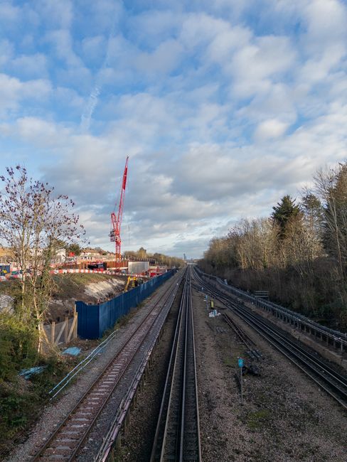 A view of multiple railway tracks extending into the distance on a clear day with partly cloudy skies. To the left, a construction site is partially enclosed by a blue temporary fence, with a tall red crane visible above. Adjacent to the site, there are sparse trees with some leaves, indicating an autumn or early winter season. On the right side, there is a line of dense trees and shrubs bordering the tracks. The tracks are accompanied by various signals and equipment, and the scene appears to be outdoors adjacent to a residential or suburban area. This image exemplifies the environment where home relocation and furniture transport services by Removal Companies Watford may involve logistical planning around railway infrastructure, especially for scheduled moving or transport of goods via rail freight.